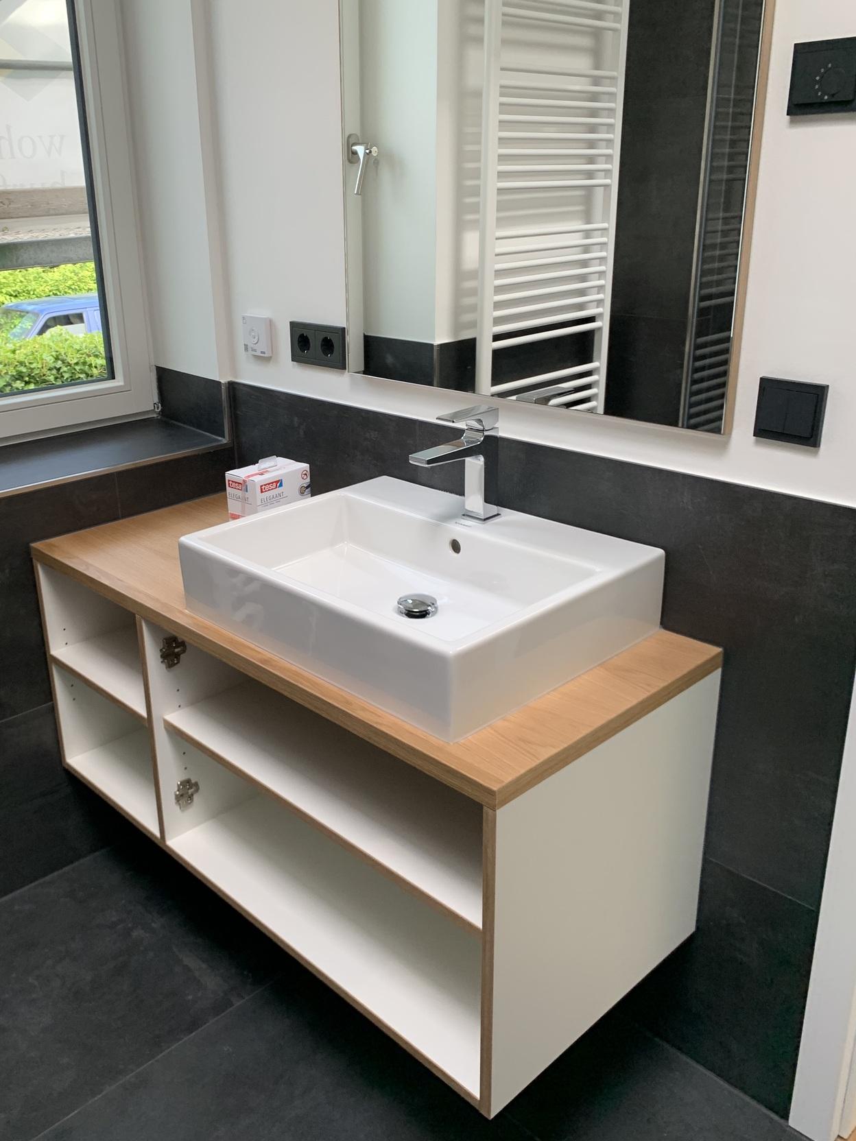 Maßgefertigter Waschtisch mit Unterschrank im Badezimmer Ein maßgefertigter Waschtisch mit weißem Waschbecken und Holzarbeitsplatte. Der offene Unterschrank bietet Stauraum. Ein großer Spiegel und ein Handtuchheizkörper ergänzen das Design. Badmöbel nach Maß.