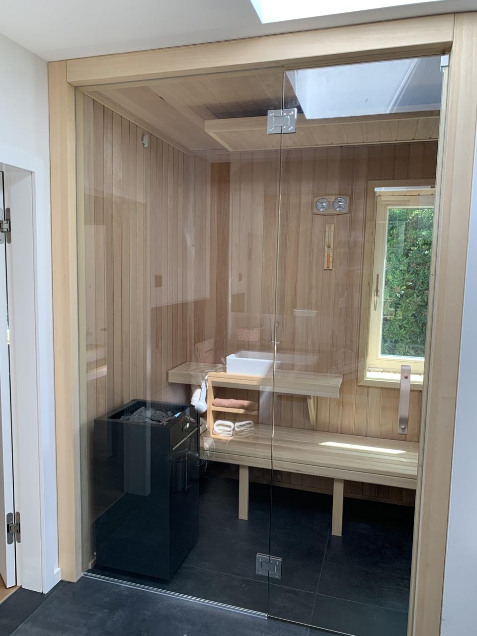 Maßgefertigte Sauna mit Glasfront und Holzinterieur Eine moderne, maßgefertigte Sauna mit Holzverkleidung, Glasfront und Sitzbank. Ein Saunaofen und ein Fenster sorgen für ein entspanntes Ambiente. Maßgefertigte Sauna.