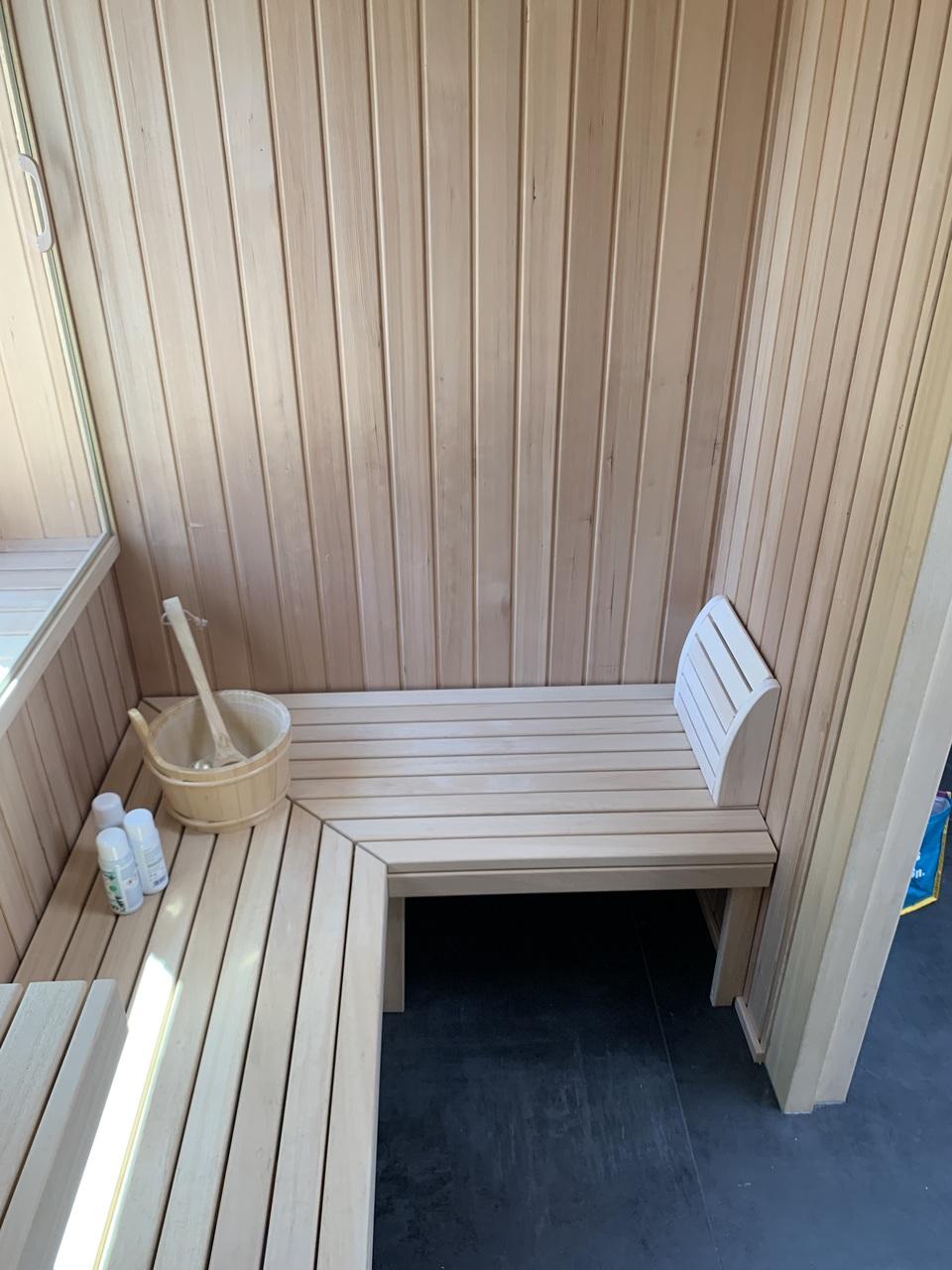 Blick von oben in eine kleine Sauna mit L-förmiger Sitzbank, holzverkleideten Wänden und Bodenrost, seitlich platzierter Saunaofen mit Schutzgitter Helle Sauna mit L-förmiger Bank, vertikaler Holzverkleidung, Rückenlehne und Fenster, dazu Saunazubehör wie Aufgusskübel und Pflegeprodukte auf der Bank