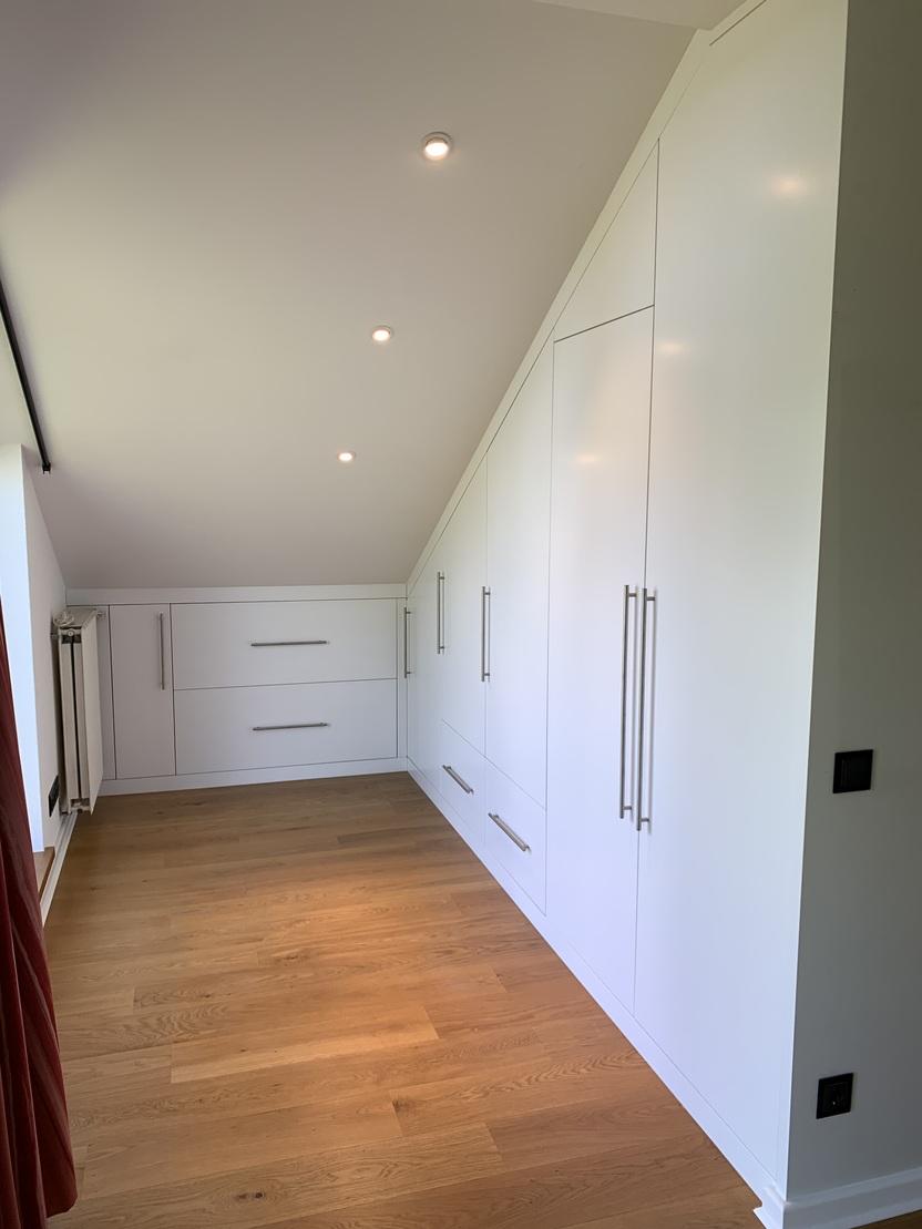 Weißer Maßschrank unter Dachschräge mit durchgehender Front Ein raumhoher Einbauschrank mit deckenbündigen weißen Fronten, Edelstahlgriffen und Schubladenauszügen, exakt an die Dachschräge angepasst, auf geöltem Eichenparkett