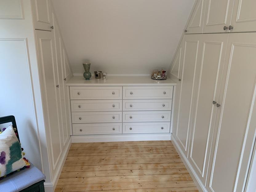 Einbauschränke mit Kommode unter Dachschräge Weiß lackierte Einbauschränke mit klassischem Kassettenprofil und mittiger Kommode mit zehn Schubladen, maßgefertigt für einen Raum mit Dachschräge und hellem Holzboden