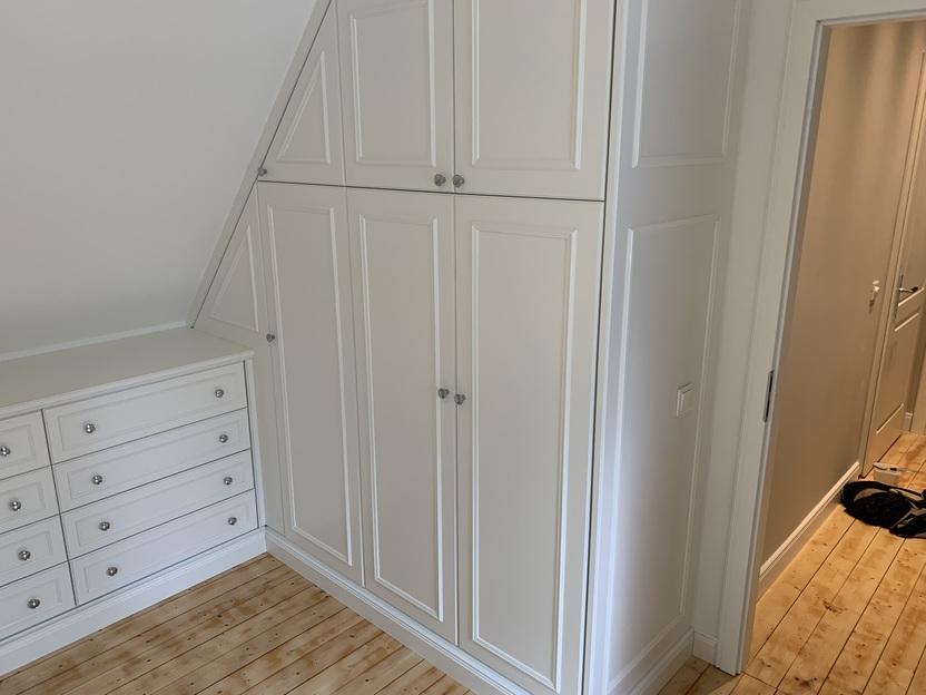 Maßgefertigter Dachschrägenschrank Ein helles Zimmer mit maßgefertigten, weißen Schränken, die an eine Dachschräge angepasst sind.