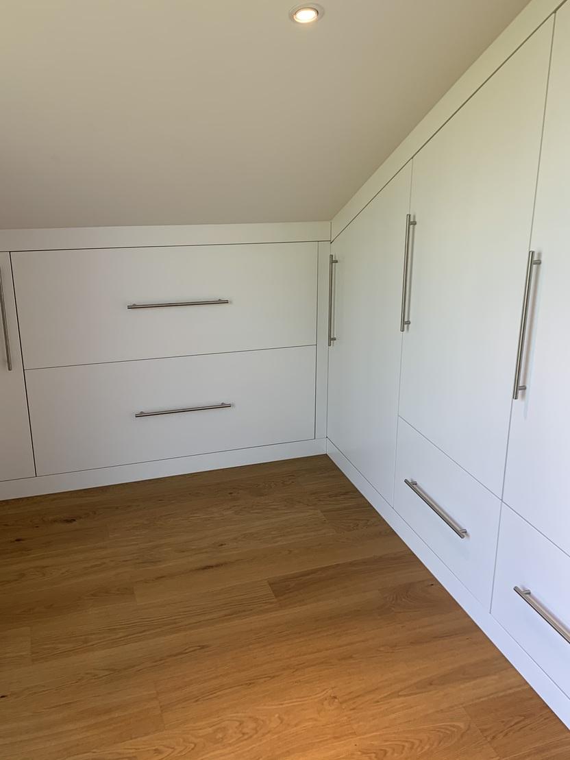 Einbauschrank unter Dachschräge mit weißer Front Individuell gefertigter Wandschrank mit glatten, weißen Fronten und Edelstahlgriffen, angepasst an eine Dachschräge, mit integriertem Heizkörperausschnitt und großem Stauraum