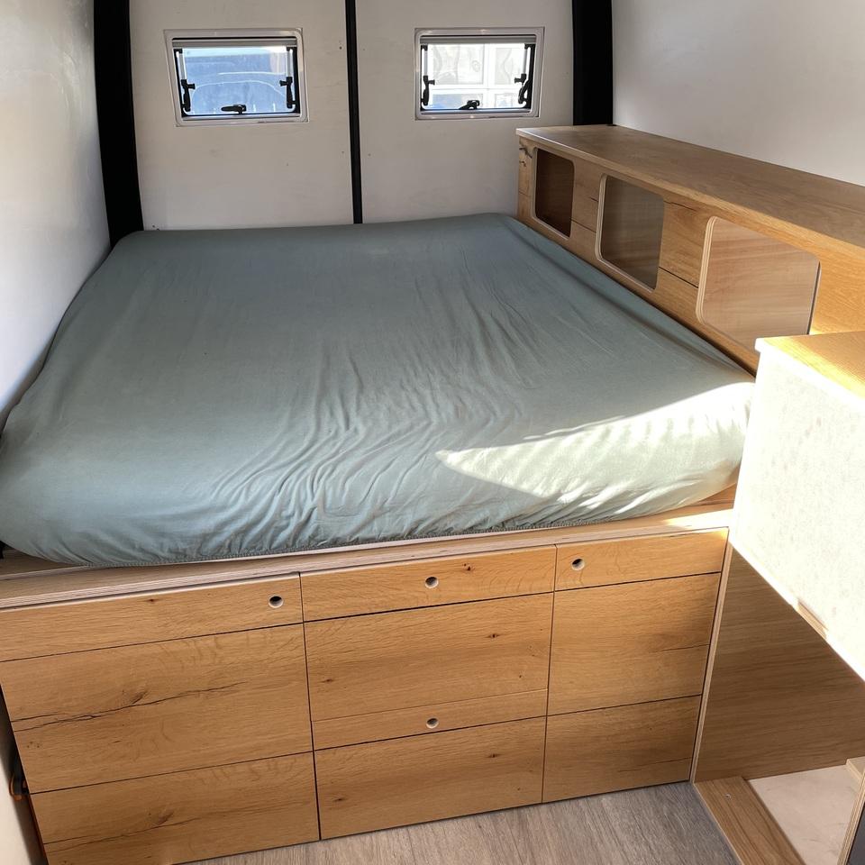 Bettmodul mit Stauraum im Wohnmobilausbau Hochwertig gefertigtes Bett mit integrierten Schubladen im Sockel und Oberschrank aus Eiche, eingebaut in einen Camper mit zwei kleinen Fensteröffnungen an der Rückwand