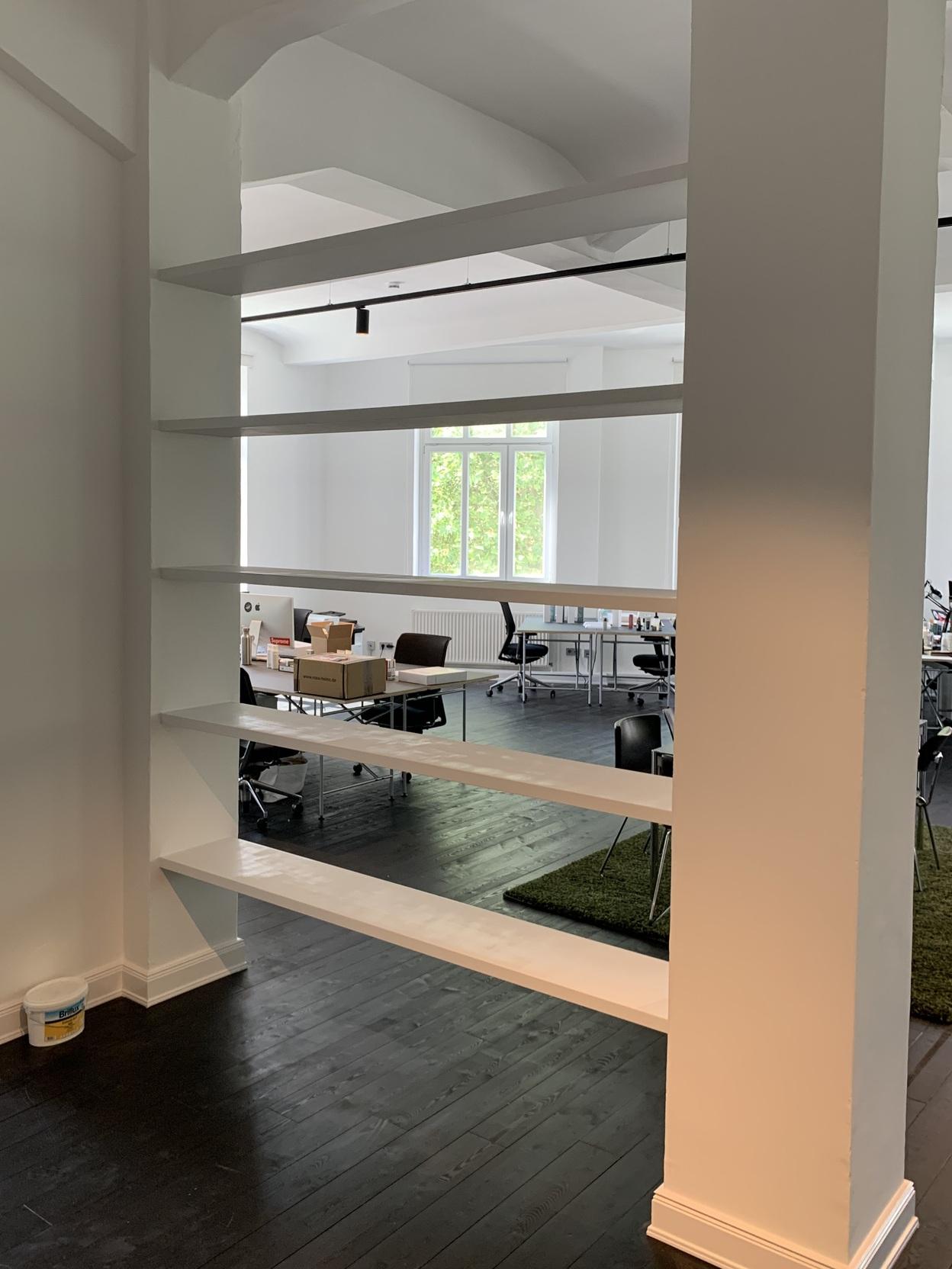 Maßgefertigtes offenes Regal als Raumteiler Ein maßgefertigtes, weißes Regal dient als offener Raumteiler in einem Bürobereich. Die schwebenden Regalbretter fügen sich elegant in die Architektur ein. Möbel nach Maß.