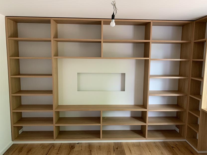 Maßgefertigte Regalwand mit integriertem Fach Eine maßgefertigte Regalwand aus Holz mit offenen Fächern und einem zentralen Fach für Dekoration oder Technik. Das Design integriert sich nahtlos in den Raum. Möbel nach Maß.