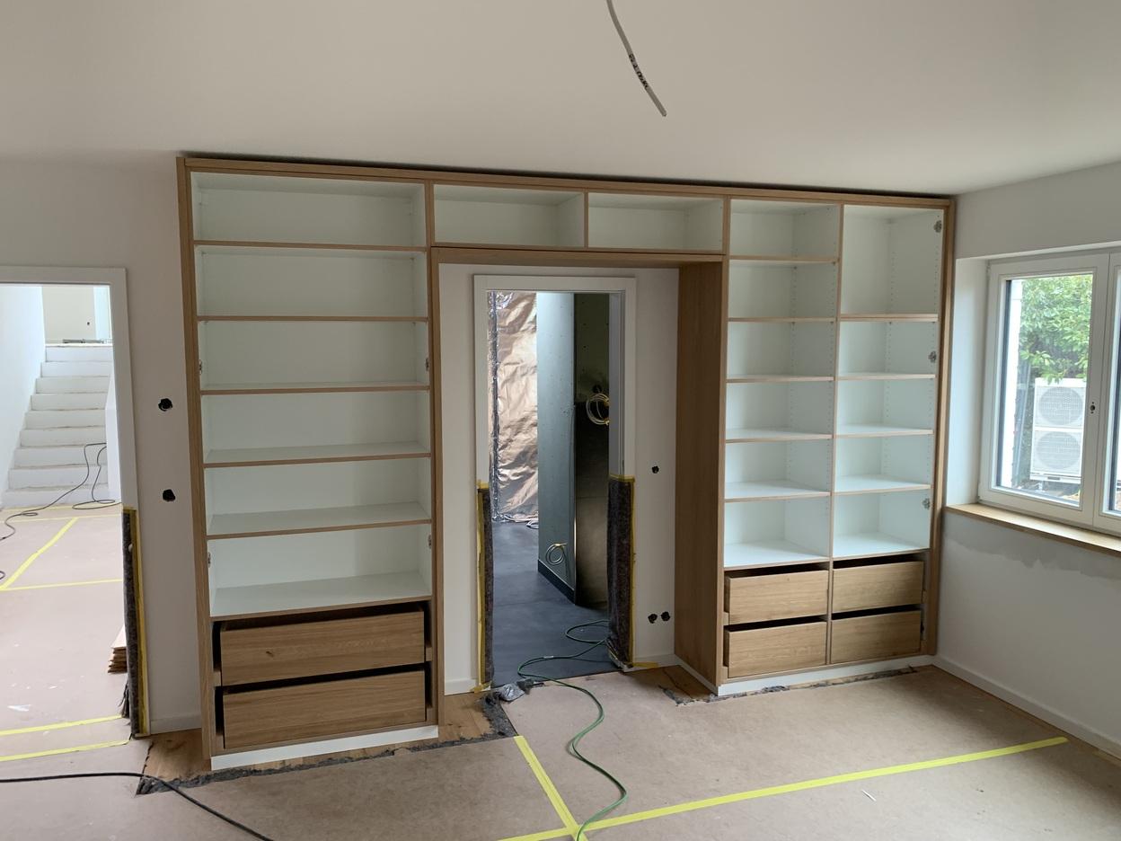 Maßgefertigter Einbauschrank mit Regalen in einem Wohnraum Ein maßgefertigter Einbauschrank aus Holz mit offenen Regalen und Schubladen, integriert in eine Wand um eine Tür herum. Der Schrank befindet sich in der Bauphase. Möbel nach Maß.