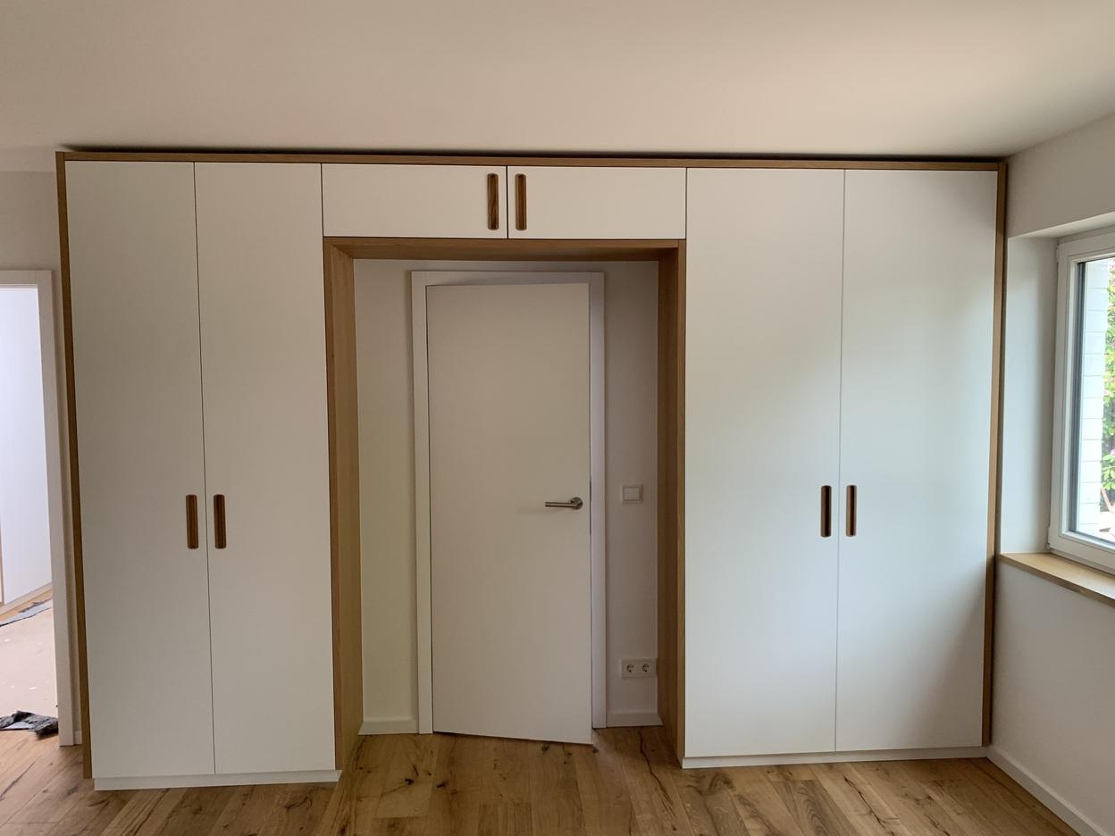 Maßgefertigter Einbauschrank um eine Tür Ein maßgefertigter Einbauschrank in einem modernen Design, der eine Tür umrahmt. Der Schrank hat weiße Fronten mit Holzgriffen und passt perfekt in den Raum. Möbel nach Maß.