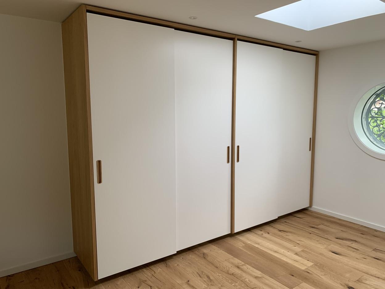 Einbauschrank mit Schiebetüren in Weiß und Holz Großer, maßgefertigter Kleiderschrank mit drei weißen Schiebetüren, seitlichen Griffausfräsungen und naturbelassenem Holzrahmen, eingebaut unter einer Dachschräge mit Oberlicht