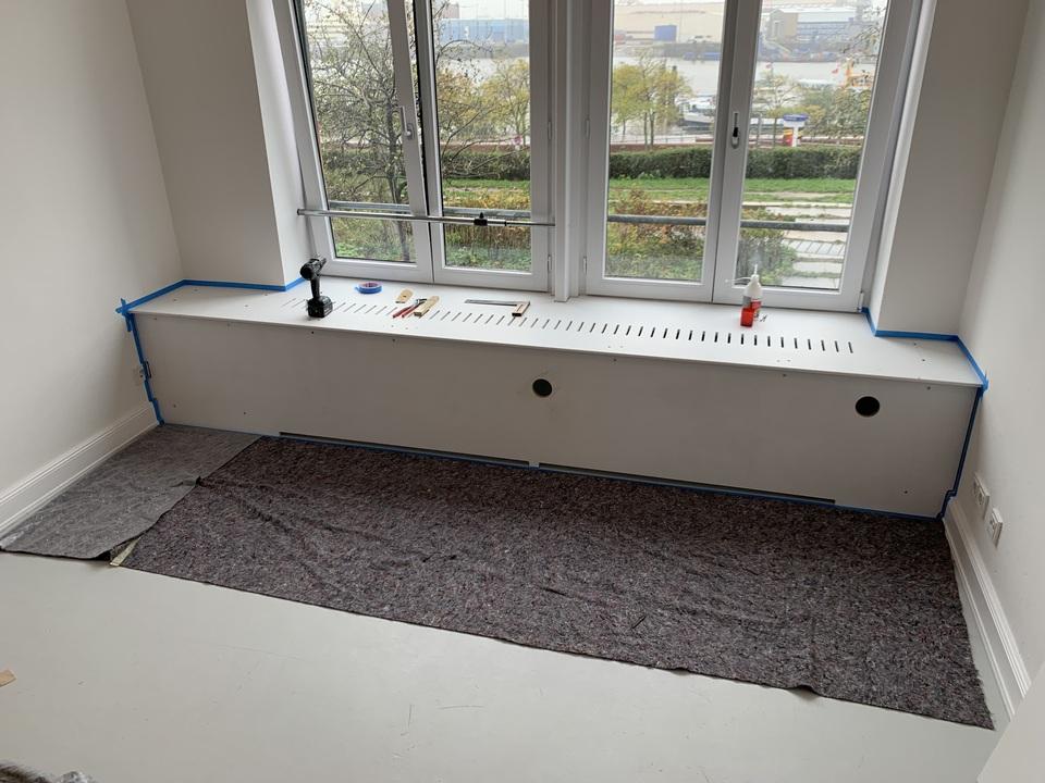 Maßgefertigte Heizungsverkleidung unter einem Fenster Eine maßgefertigte Heizungsverkleidung wird unter einem großen Fenster montiert. Das Möbelstück integriert sich nahtlos in den Raum. Werkzeuge sind für die Fertigstellung bereit. Möbel nach Maß.