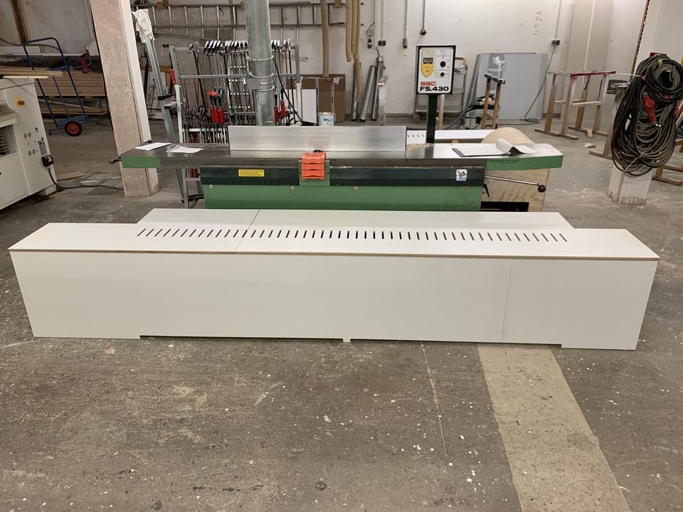 Maßgefertigte Heizungsverkleidung mit Luftschlitzen Weiße Verkleidung mit schmalen Lüftungsschlitzen auf der Oberseite, in der Werkstatt vor einer Maschine aufgestellt, gefertigt als Maßarbeit