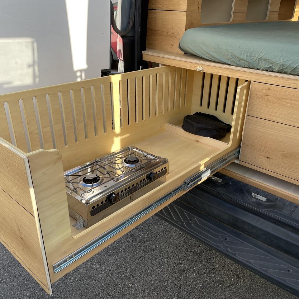 Ausziehbare Gasküche im Camperausbau Seitlich ausziehbares Küchenmodul mit zweiflammigem Gaskocher, belüfteter Rückwand und Holzverkleidung, integriert unter dem Bett im selbst ausgebauten Wohnmobil