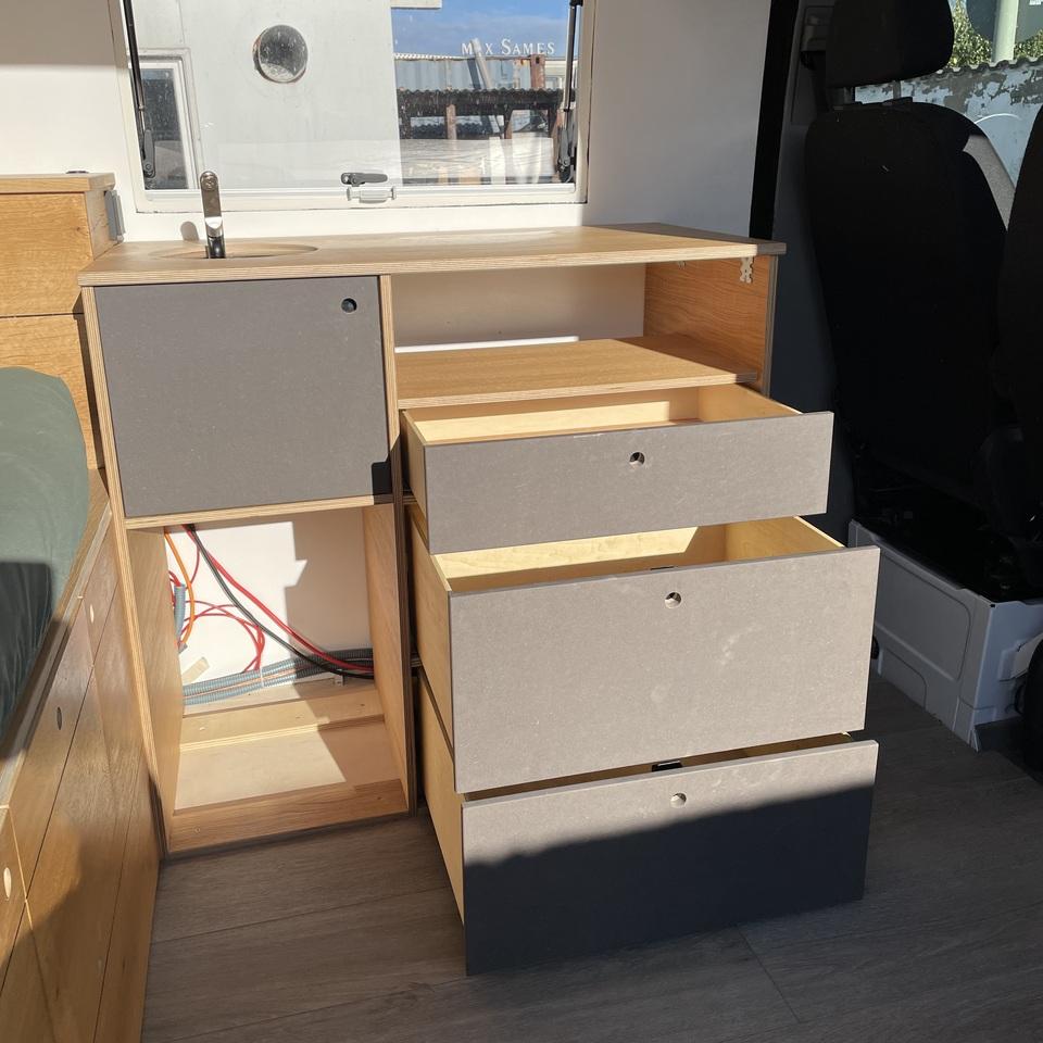Offene Schubladenelemente im Camperausbau Camperausbau mit drei geöffneten Schubladen in anthrazitfarbenen Fronten, integriertem Technikfach mit sichtbarer Verkabelung und Spüle unter dem Fenster