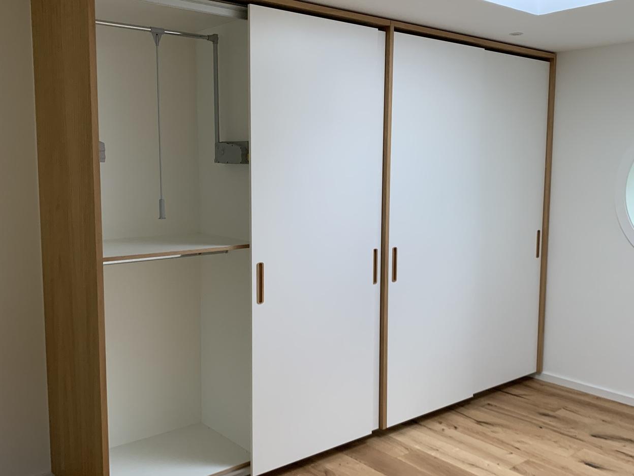 Schiebetürschrank mit offenem Fach und Kleiderlift Maßgefertigter Schrank mit drei weißen Schiebetüren und geöffneter Abteilung links, darin ein ausziehbarer Kleiderlift und eine Kleiderstange, kombiniert mit hellem Holzdekor und Griffmulden